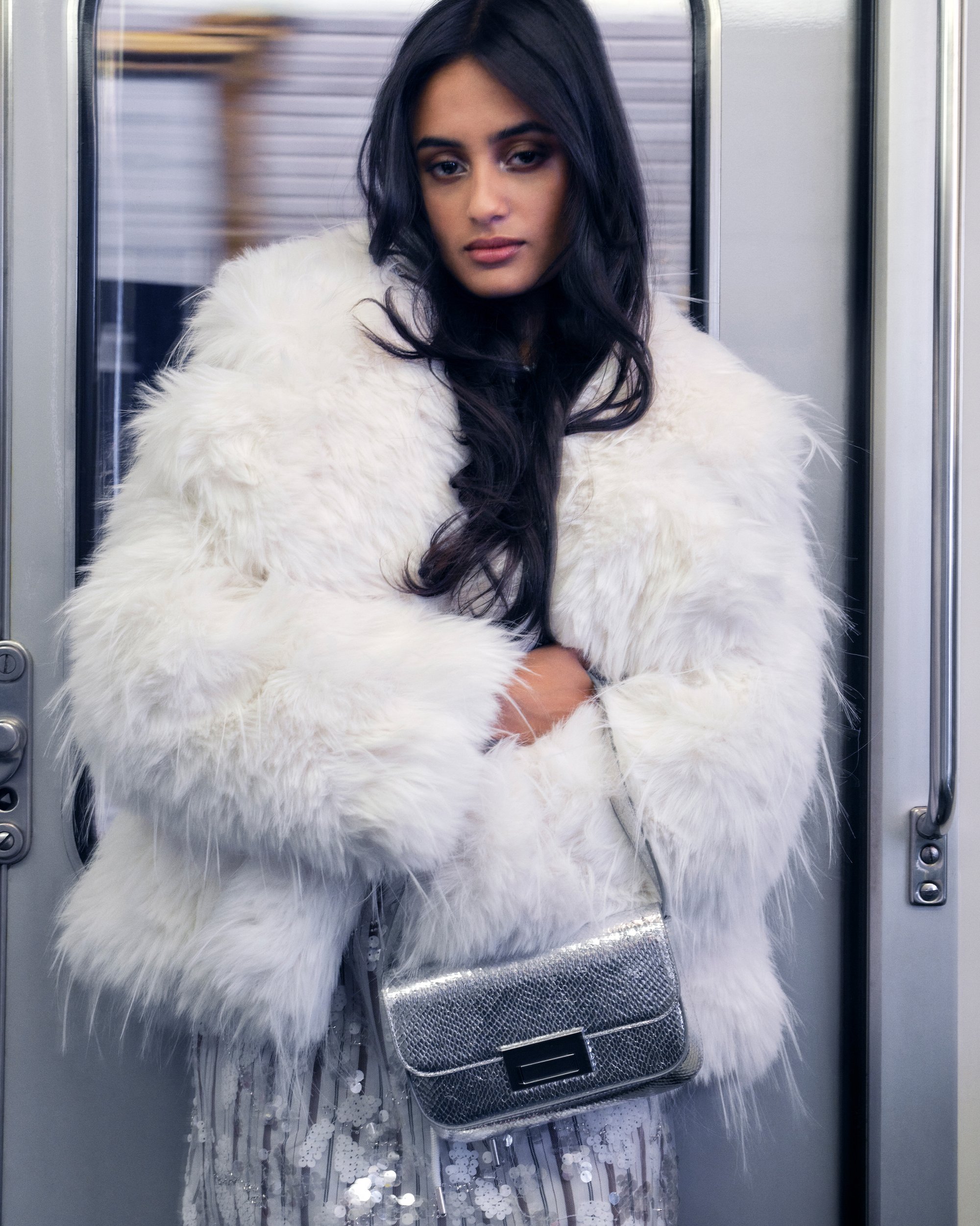 Een vrouw in een wit bontjas staat in een metrotrein, houdt een zilveren handtas vast en kijkt nadenkend, omringd door grijze interieur elementen.