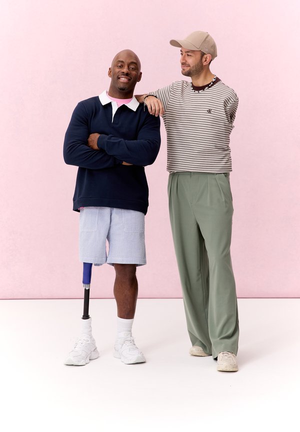 Zwei Männer stehen vor einem rosa Hintergrund; einer mit Prothesenbein trägt Shorts und Sweatshirt, der andere ein gestreiftes Hemd und grüne Hosen.
