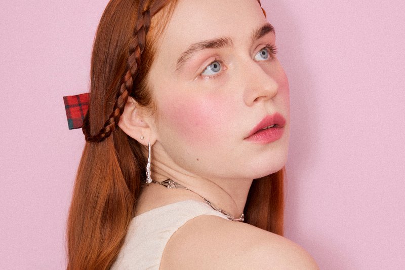 Young woman with long red hair braided and tied with a red plaid ribbon, wearing silver earrings and a light sleeveless top against a pink background.