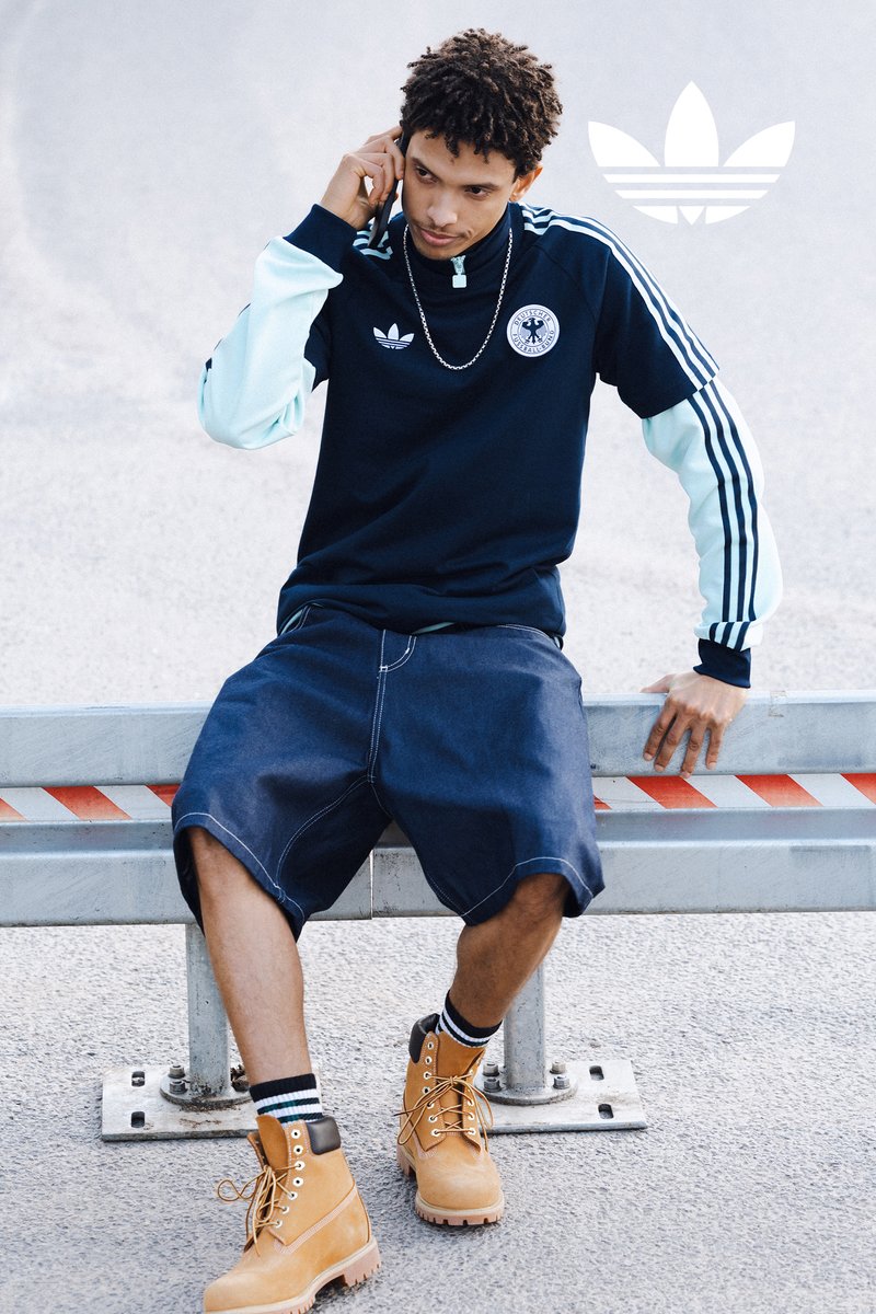 Junger Mann in Sportbekleidung, der auf einer Metallbarriere sitzt, telefoniert und eine blaue Adidas-Jacke, Shorts, gestreifte Socken und beige Stiefel trägt.