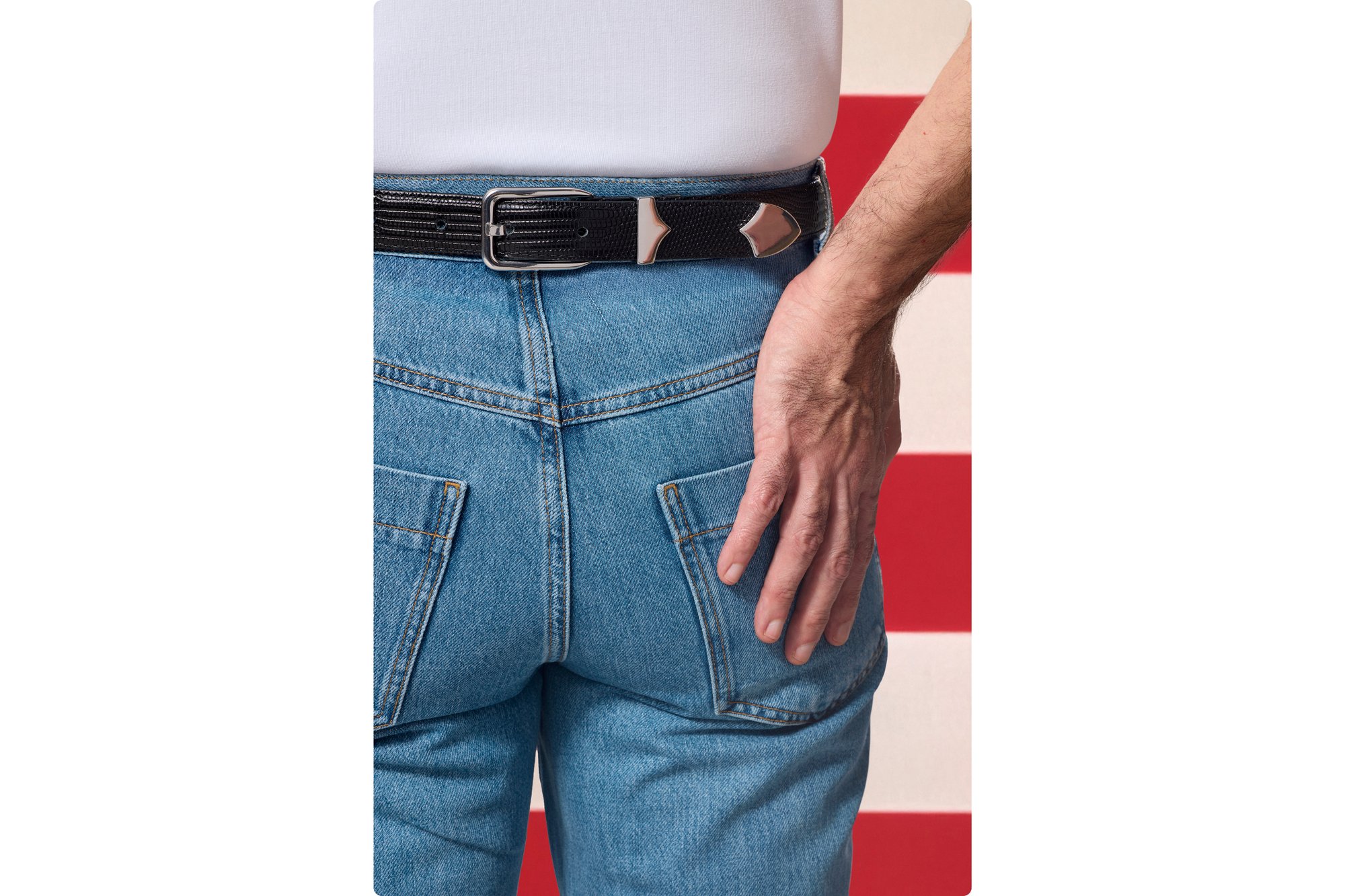 Una persona è in piedi con le spalle alla camera, indossando una camicia bianca e jeans blu, con la mano appoggiata all'anca, su uno sfondo a strisce.