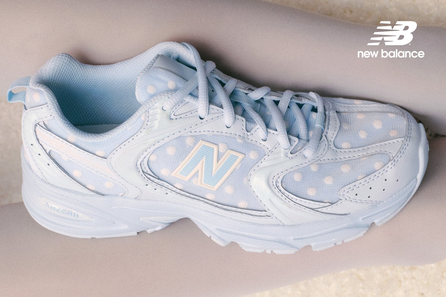 Sneakers azzurri con pois bianchi, caratterizzati da un grande logo "N", poggiati su una superficie morbida. Stile elegante e minimalista.