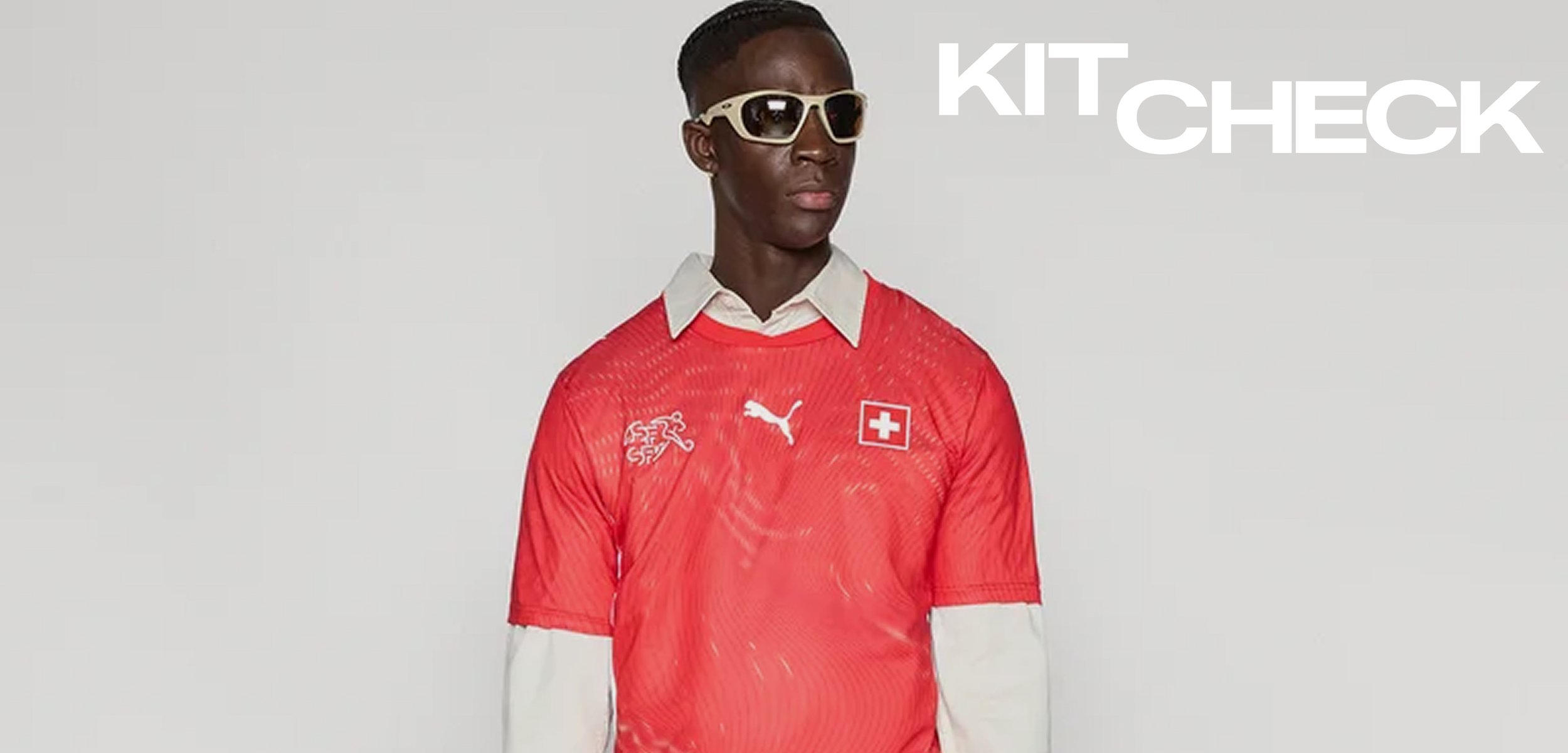 Bărbat purtând tricou de fotbal elvețian roșu cu logo Puma alb și ochelari de soare, stând în fața unui fundal simplu cu textul "KIT CHECK".