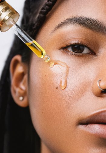 Une huile pour le visage claire est gouttée sur la peau, de couleur jaune doré. Texture lisse sur la peau, mettant en avant un éclat naturel. Un œil brun et des cheveux tressés sont visibles.