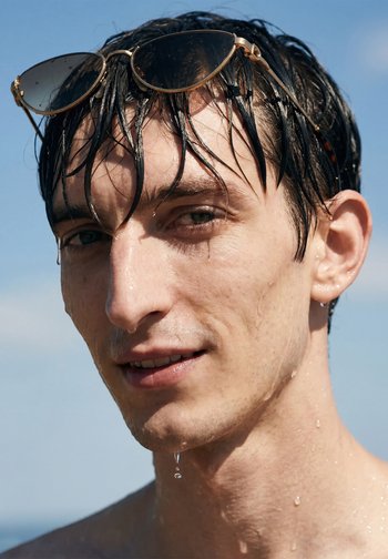 Jeune homme aux cheveux mouillés et lunettes de soleil sur la tête, gouttelettes d'eau sur le visage et le cou, sur un fond de ciel bleu clair.