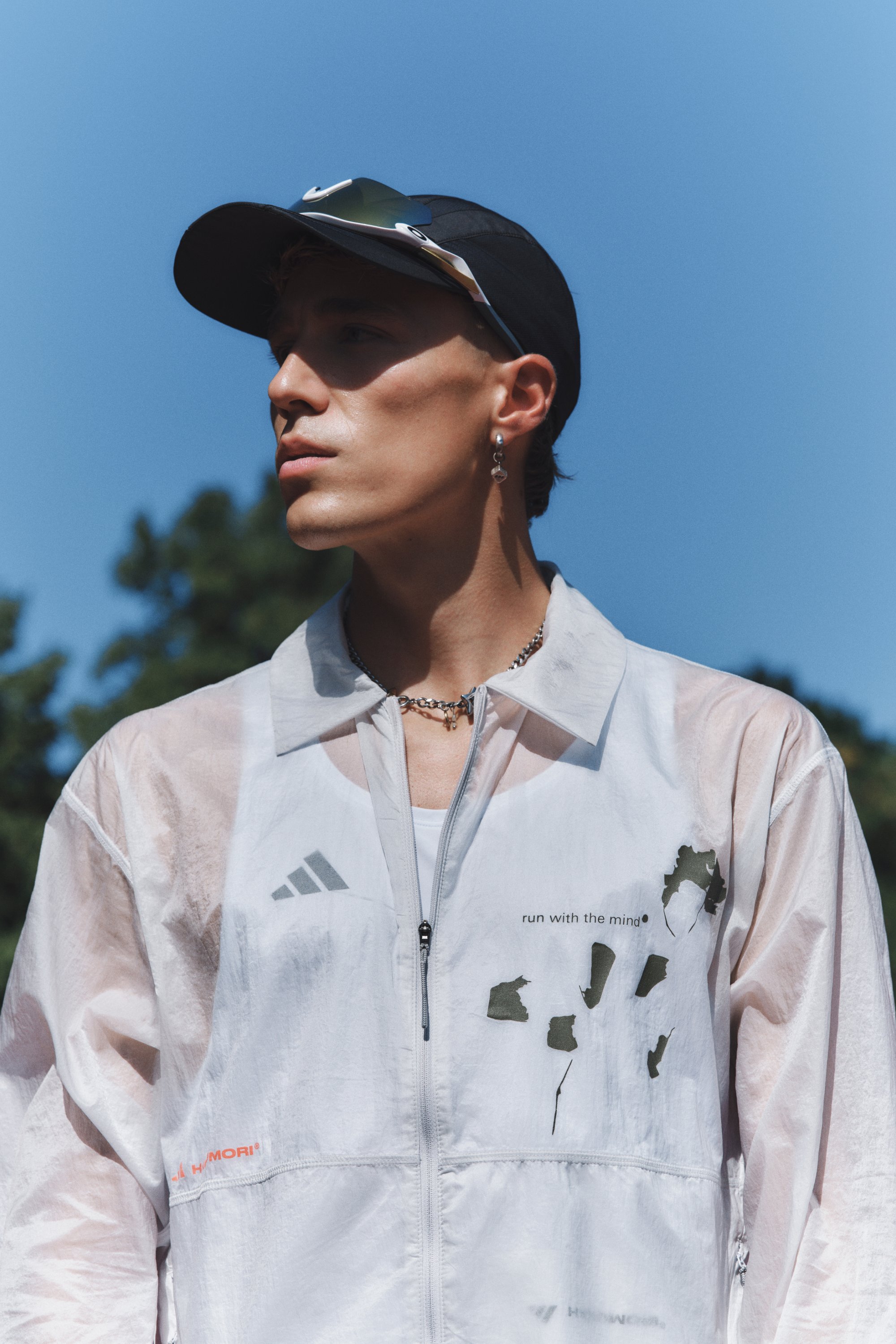 Lichtgewicht, doorzichtig wit jack met ritssluiting aan de voorkant, Adidas-logo en groen abstract patroon. Draag het over een wit tanktop, gecombineerd met een zwarte pet en een zonnebril.