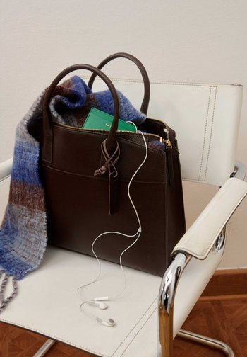 Bolso de mano de cuero marrón oscuro sobre una silla blanca con una bufanda azul marrón, cartera verde en el interior y auriculares blancos colgando por delante.
