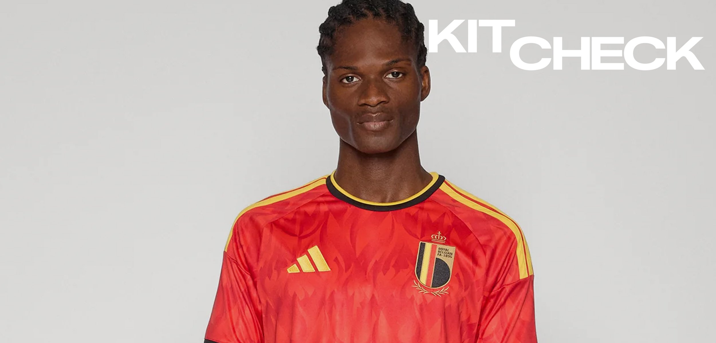 Homme portant le maillot rouge de l'équipe nationale belge de football avec des rayures jaunes et un col noir, debout devant un fond uni.
