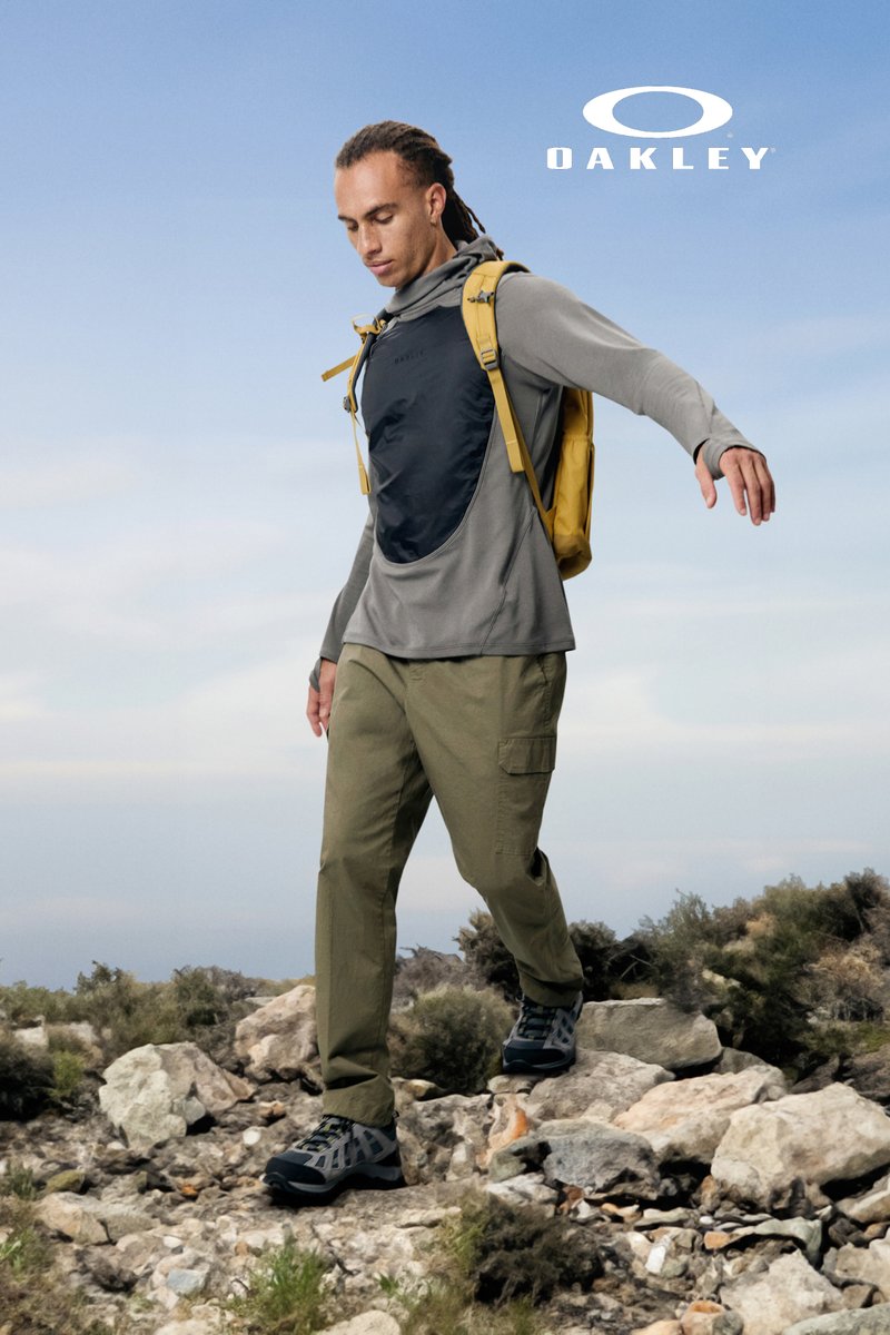 Una persona con el pelo largo, vistiendo una camiseta gris y pantalones cargo, camina sobre terreno rocoso, llevando una mochila amarilla, contra un cielo azul.