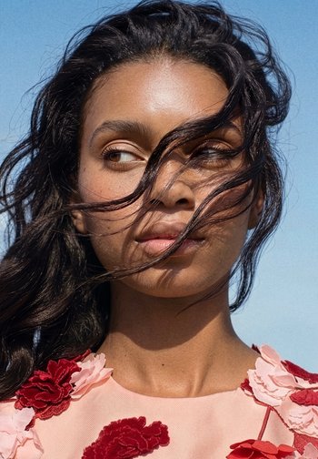 Young woman with long dark hair blowing across her face, wearing a pink top adorned with red and pink floral appliqués, against a clear blue sky.