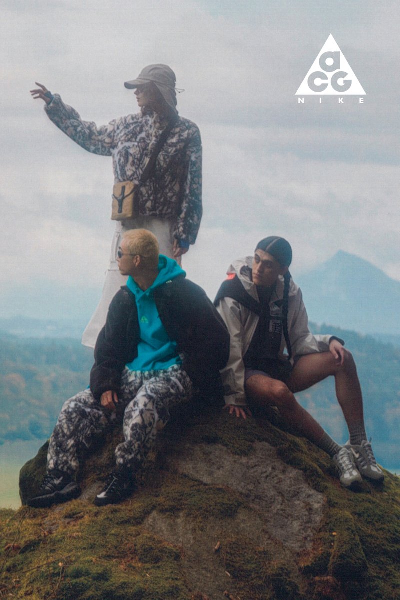 Tres jóvenes posan sobre una roca al aire libre. Uno señala mientras los otros dos están sentados, vestidos con ropa de exterior casual y a la moda, frente a un nebuloso fondo montañoso.