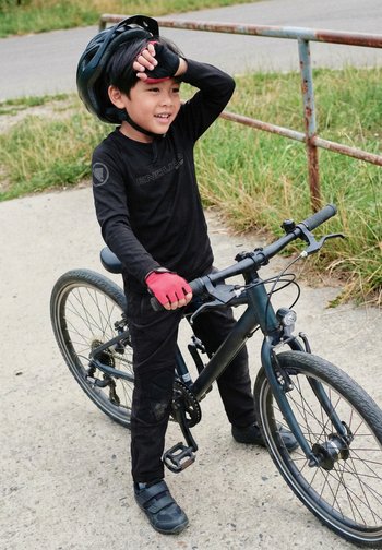 Junger Junge in schwarzer Radbekleidung und Helm steht auf einem befestigten Weg nahe Gras und hält ein schwarzes Fahrrad.