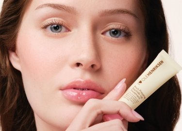 Tubka kremu w jasnobeżowym opakowaniu z czarnym nadrukiem; modelka o jasnej karnacji, czystej cerze i naturalnym kolorze ust. Skóra wygląda na nawilżoną.