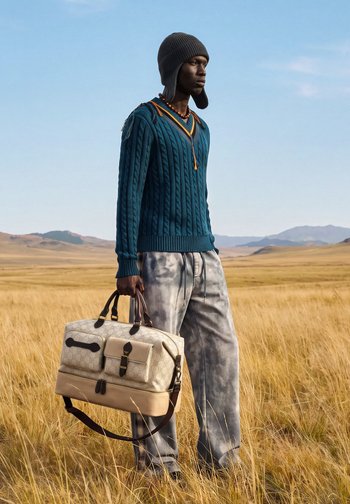 Homme portant un chapeau gris, un pull torsadé bleu sarcelle et un pantalon tie-dye tenant un grand sac beige de créateur dans un champ d'herbe sèche avec des montagnes au loin.
