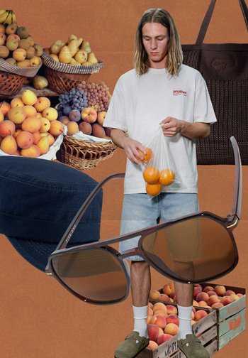 Jeune homme en chemise blanche et short tenant un sac d’oranges, entouré de paniers de fruits, lunettes de soleil, casquette marine et sac marron.