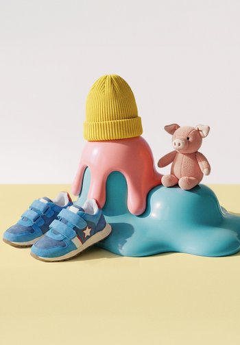 Blaue Kindersneaker, rosa Kuschelschwein-Spielzeug, gelbe Strickmütze auf einer rosa-blauen abstrakten Skulptur vor einem hellen Hintergrund.