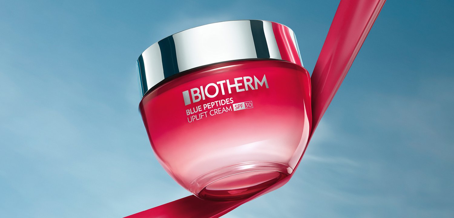 Pot rouge de crème Uplift de Biotherm avec peptides bleus et protection solaire SPF 30, suspendu contre un ciel bleu avec un ruban rose flotter.