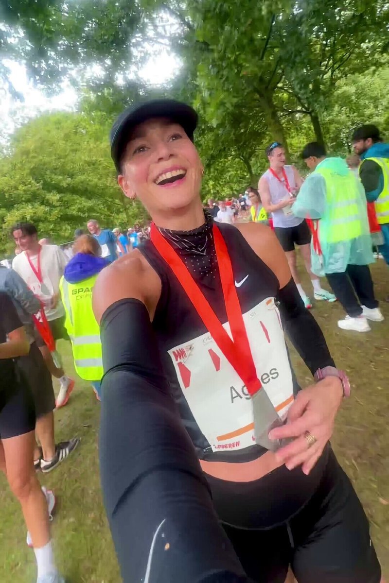 Osoba oblečená v černém sportovním tílku a legínách drží medaili s červenou stužkou. Je viditelný jmenovka. Scénu rámují zelené stromy.