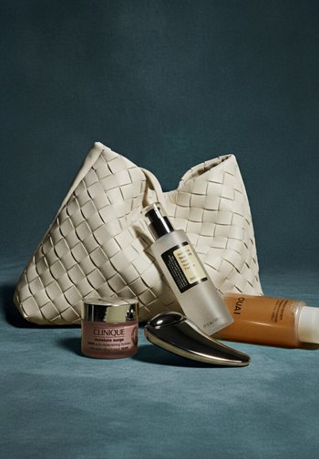 Bolso blanco tejido, botella de suero transparente, tarro de crema rosa, aplicador de metal y botella de champú naranja sobre un fondo azul texturizado.