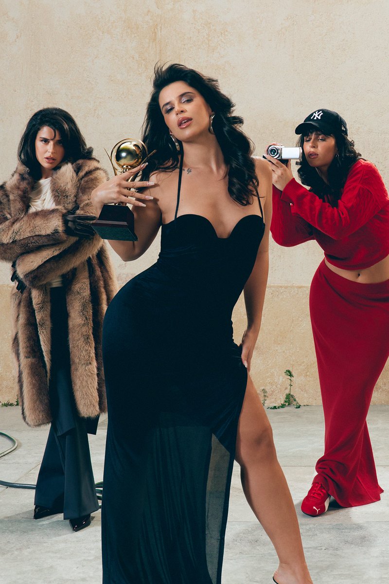 Tres mujeres posan en un estudio. Una lleva un vestido negro y sostiene un trofeo, mientras que otra, con un abrigo de piel, está cerca. Una tercera toma una foto.