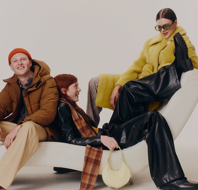Three young adults wearing winter clothes, sitting and lounging on a white modern chair, smiling and interacting.