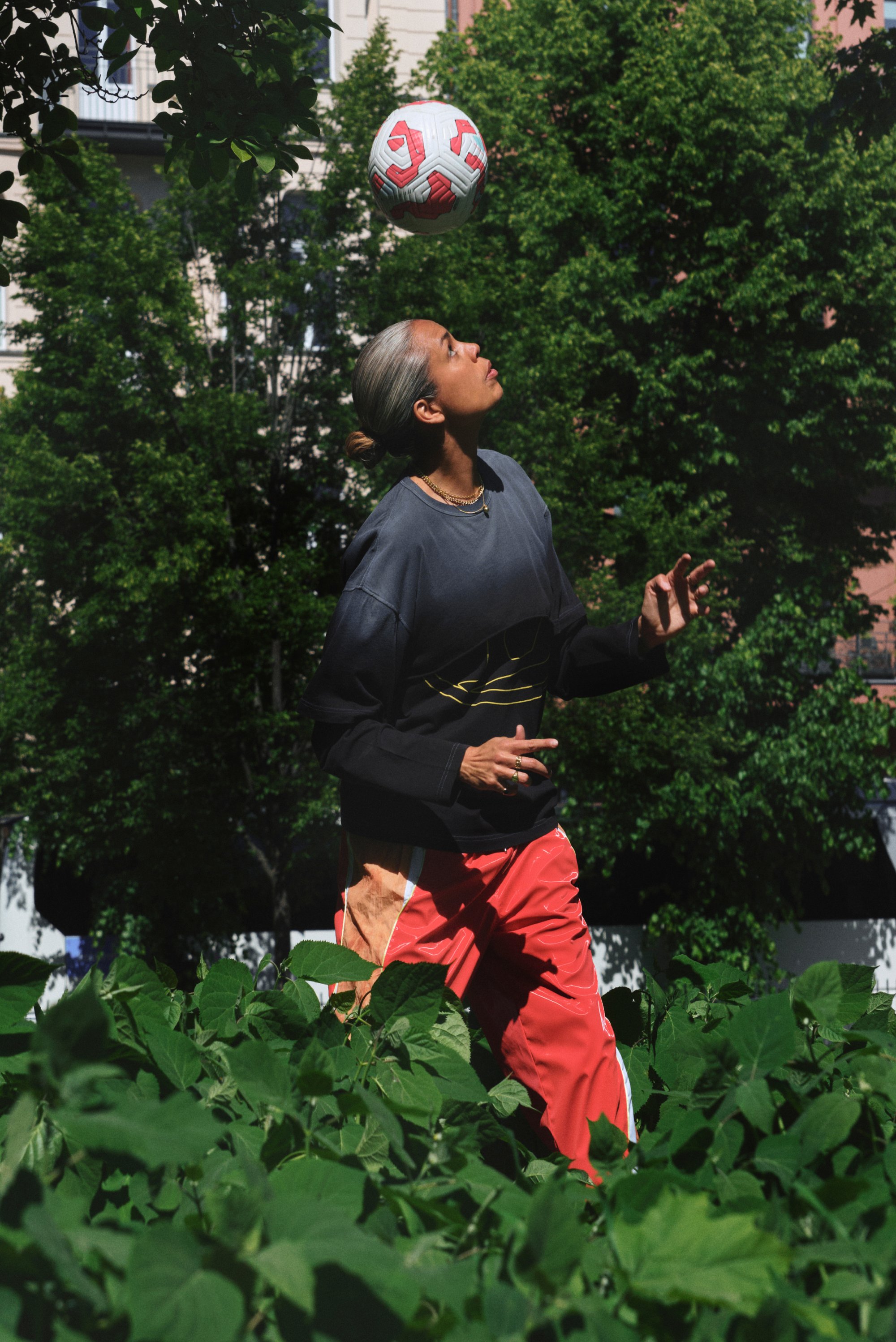 A person with long hair in a black and orange outfit is juggling a soccer ball while standing in a dense green field under sunlight.