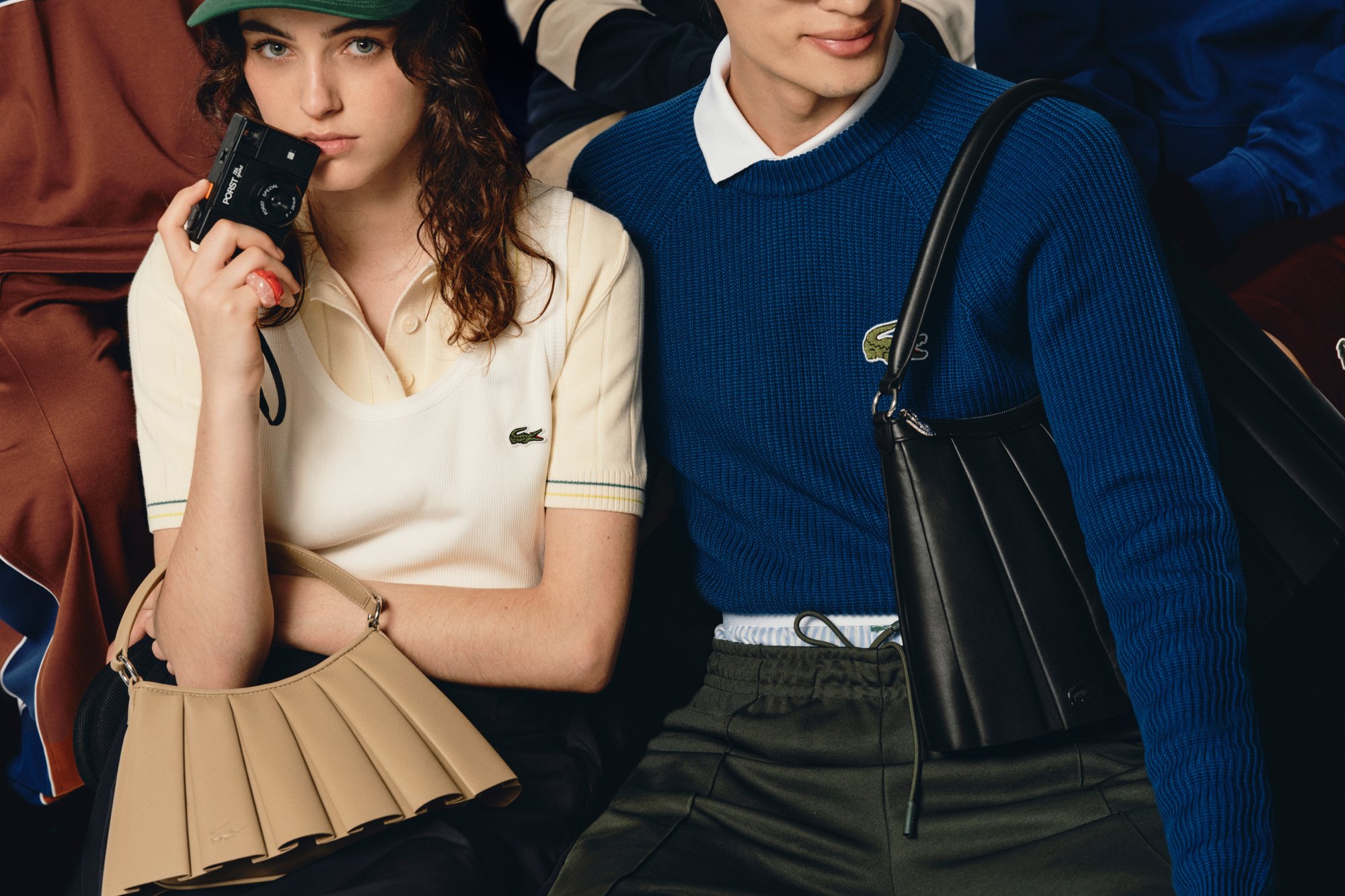 Una donna con i capelli ricci tiene una macchina fotografica, indossando una polo crema e una borsa beige. Accanto a lei, un uomo in un maglione blu mostra una borsa nera.