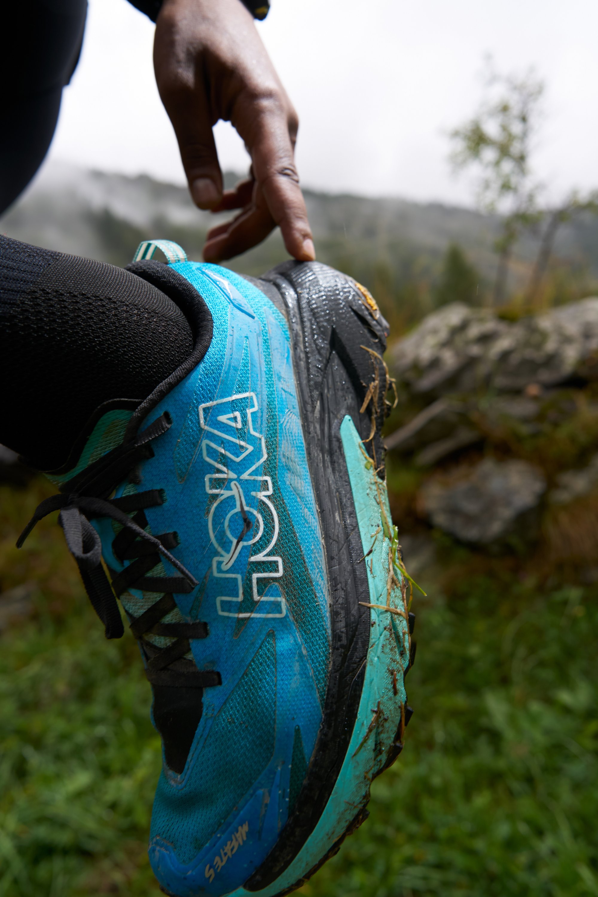 Blue and black HOKA trail running shoe featuring a textured upper, a prominent logo, and visible mud on the sole. The accent colours are teal.