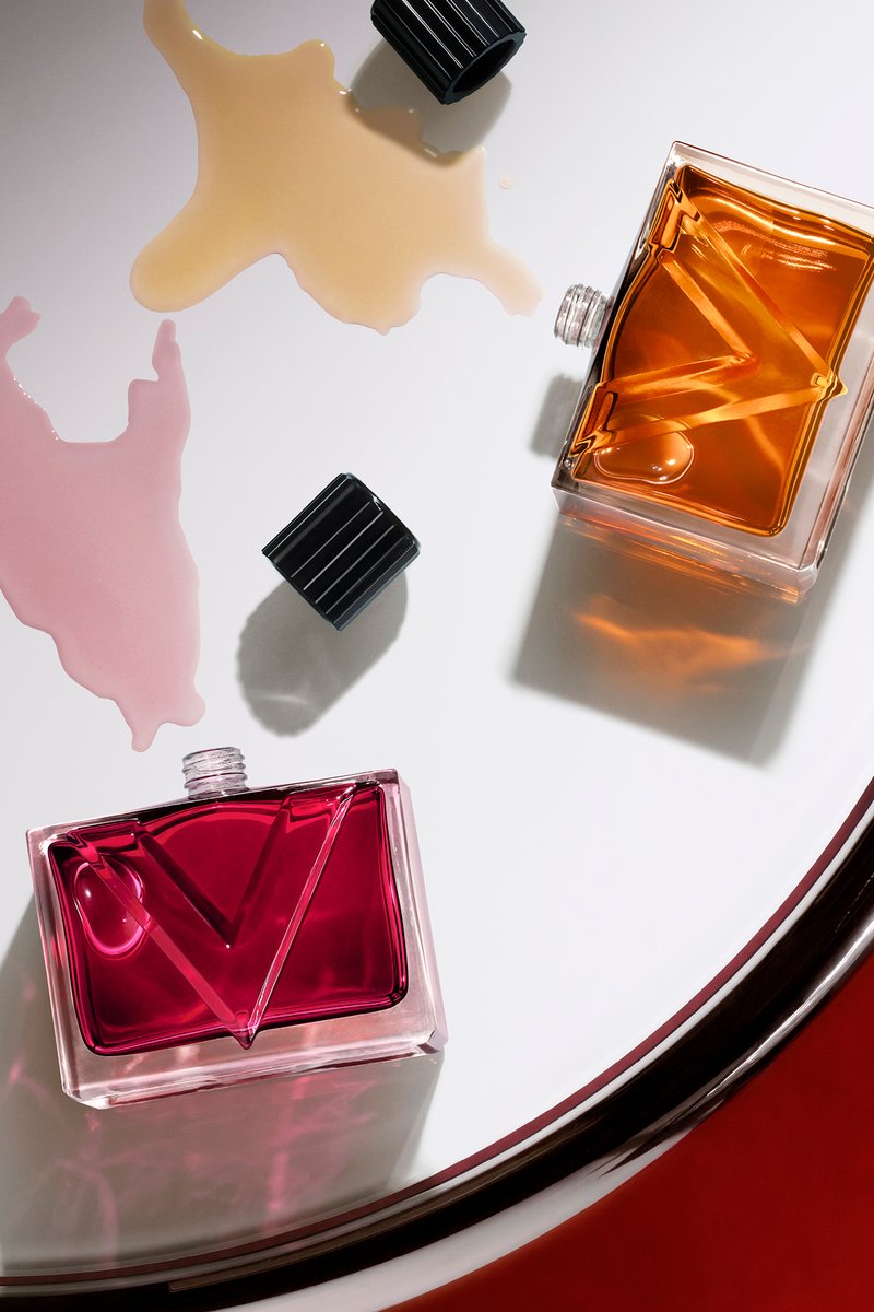 Deux flacons de parfum ouverts, un rouge et un ambré, avec du liquide renversé et des bouchons noirs sur une surface blanche ronde.