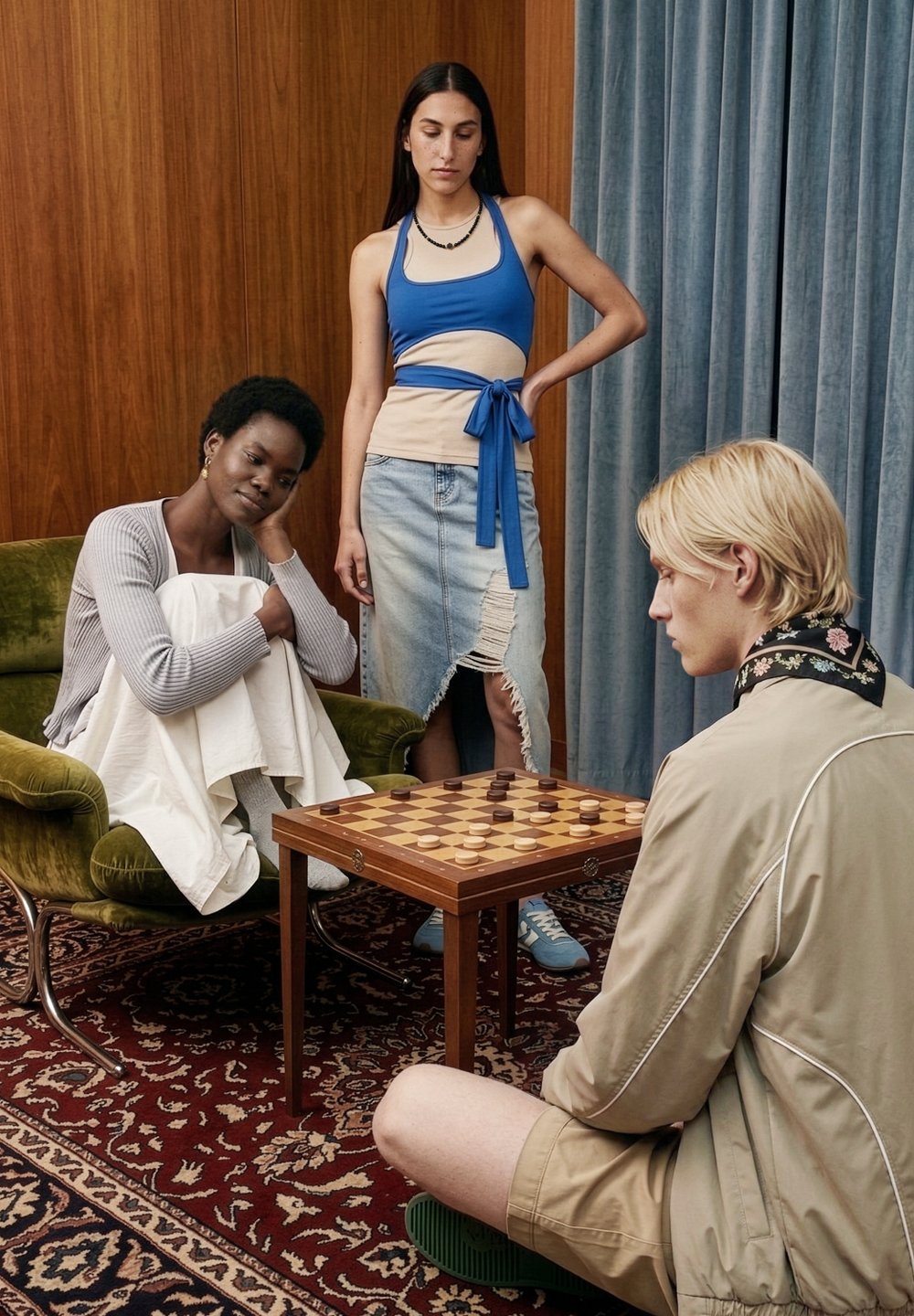 Trois personnes dans une pièce avec des rideaux en bois et bleus, deux assises et une debout, jouant aux dames sur un plateau en bois sur une petite table.
