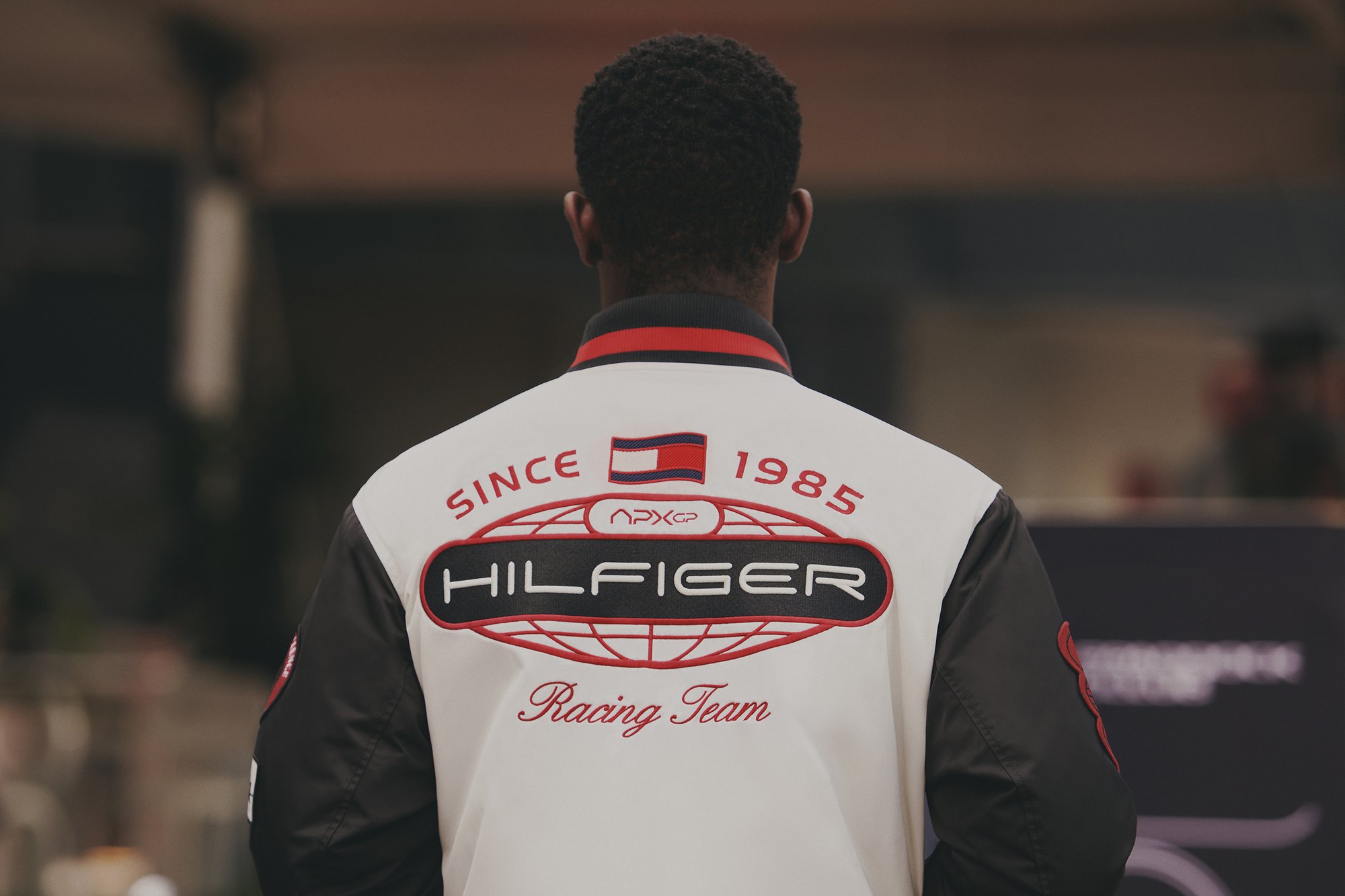 Una persona de espaldas viste una chaqueta blanca con el logo del "Hilfiger Racing Team" y el texto "Desde 1985", en un ambiente de evento casual.