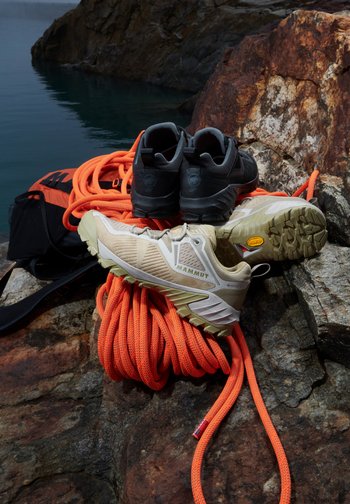 Two pairs of climbing shoes, one beige and one black, rest on vibrant orange climbing rope atop a rocky surface near water. A black backpack is partially visible.