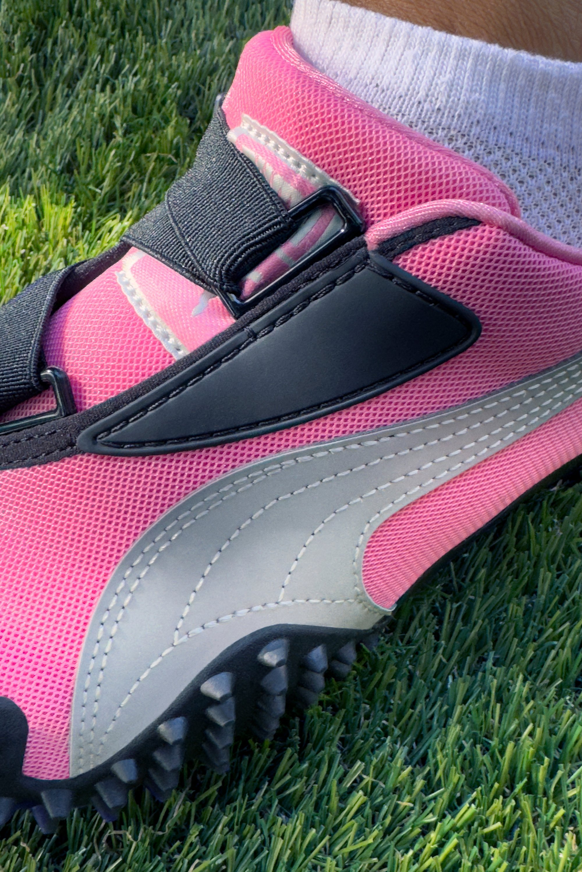 Zapatilla deportiva Puma en malla de color rosa brillante con detalles en negro, logotipo plateado y suela texturizada con prominentes tachuelas para mayor tracción.