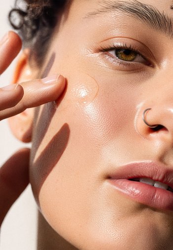 Close-up of a face showcasing smooth, glowy skin. A finger applies a clear, gel-like product near the cheek. Features include a septum ring.