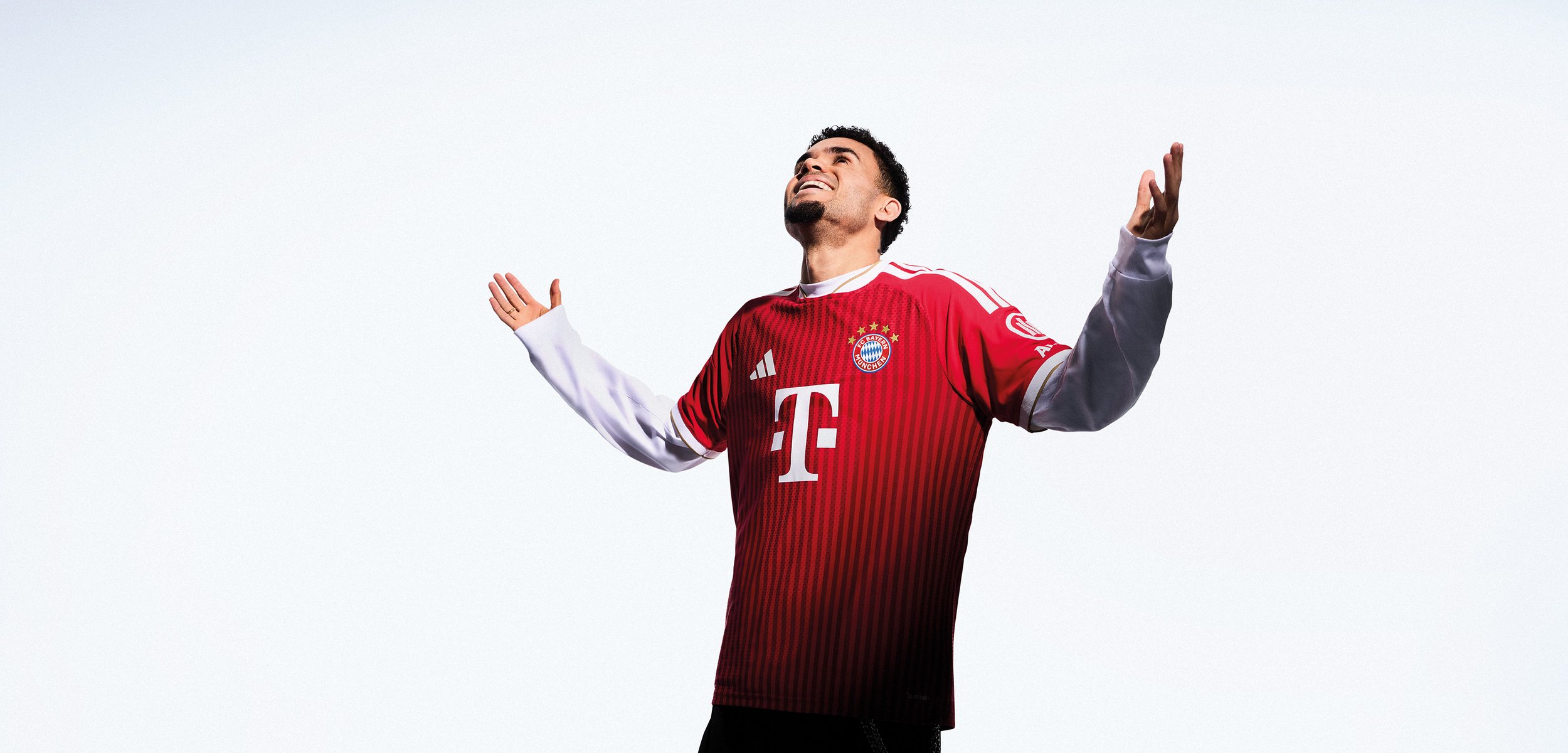 Mann im Bayern München Fußballtrikot hebt die Arme und schaut vor weißem Hintergrund nach oben.