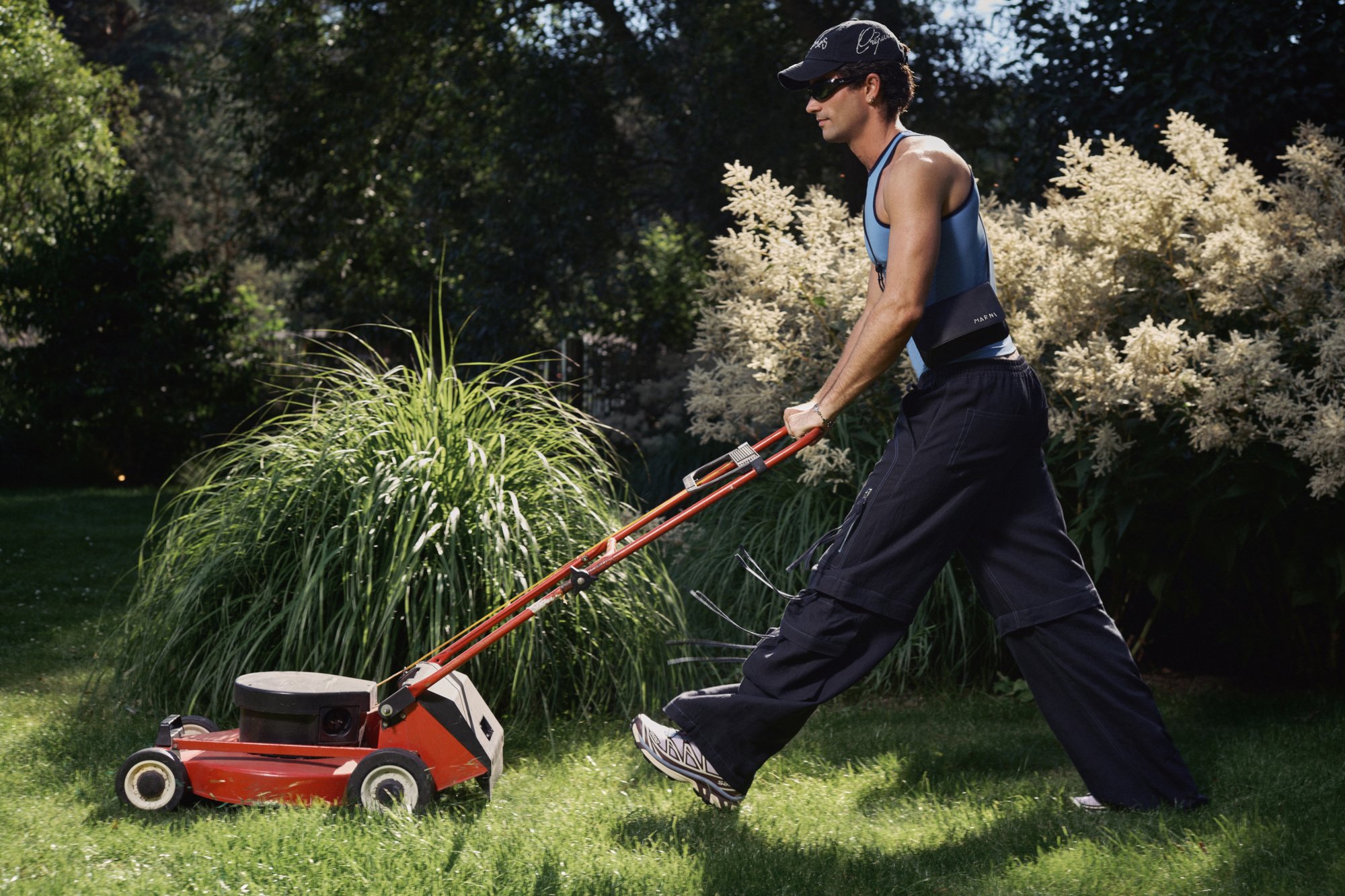 A man wearing a sleeveless shirt and cap is pushing a red lawnmower across a grassy area, encircled by tall green plants and flowers.