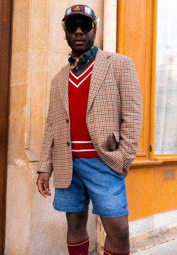 Eine Person steht an einer strukturierten Wand und trägt ein gemustertes Blazer, einen roten Pullover und Jeansshorts, ergänzt mit Sonnenbrille und einer Mütze.