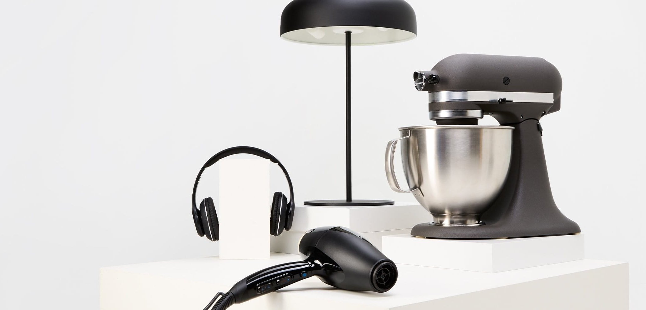 Mikser stojący w matowej czerni ze stalową misą, czarna suszarka do włosów, słuchawki i czarna lampa stołowa na białych powierzchniach wystawowych.