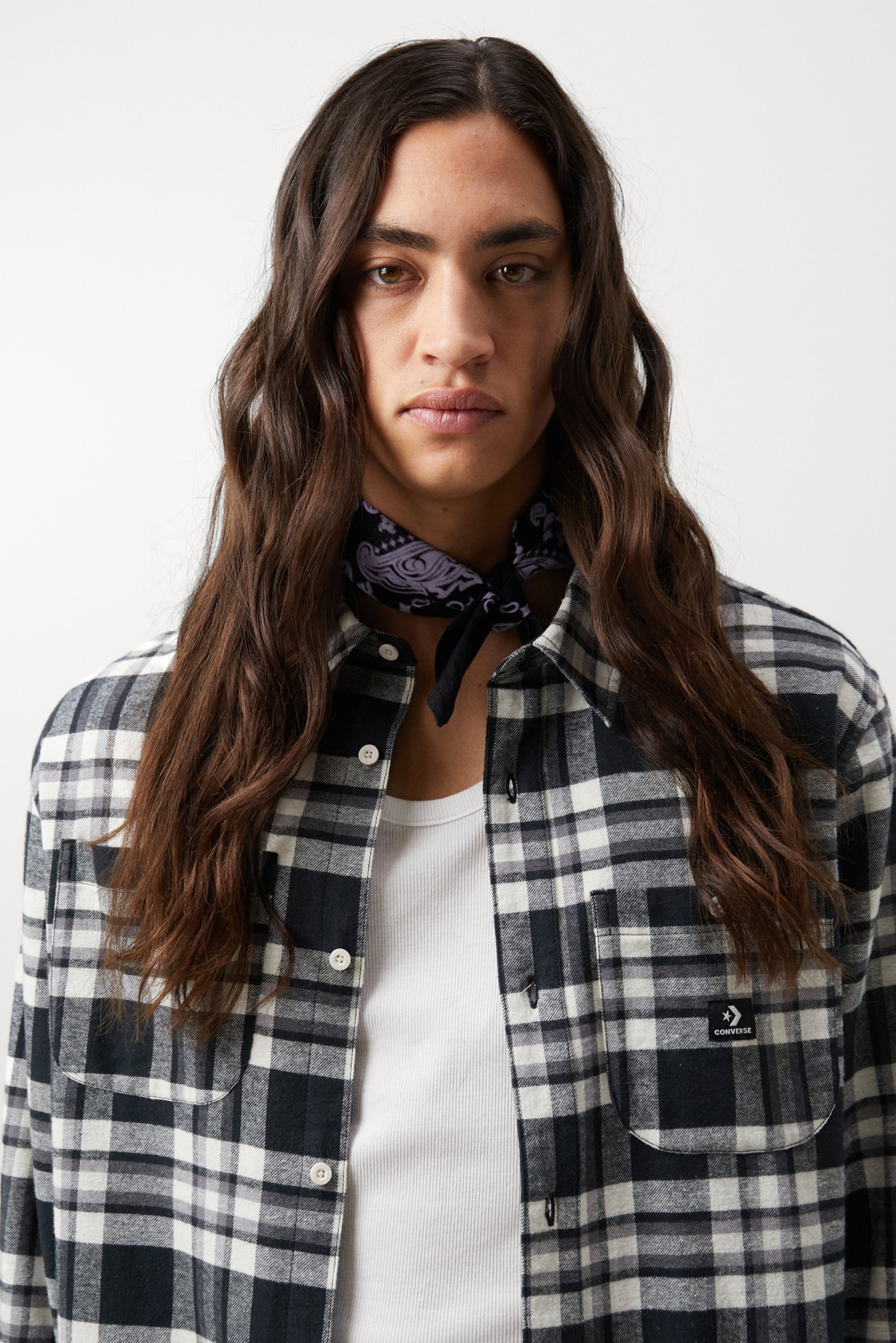 A person with long, wavy hair wears a black and white plaid shirt over a white tank top, accessorized with a black paisley scarf.