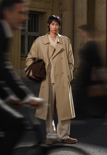 Un trench-coat beige à double boutonnage superposé sur un pull léger et un pantalon crème. Le modèle porte un sac à bandoulière marron foncé.