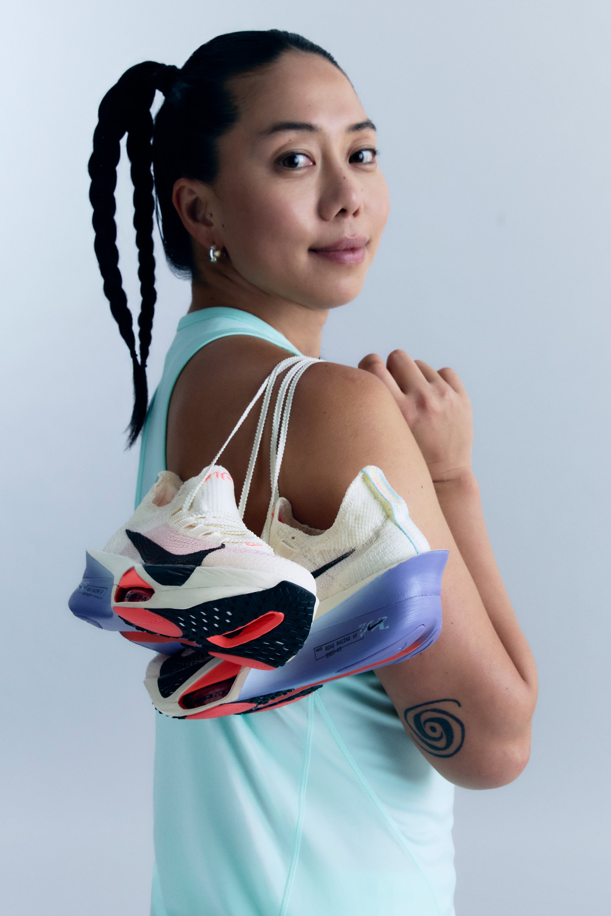 Des chaussures de course avec une tige crème ornée d'accents noirs et roses, une semelle texturée et un talon violet, tenues par une personne portant un débardeur bleu clair.