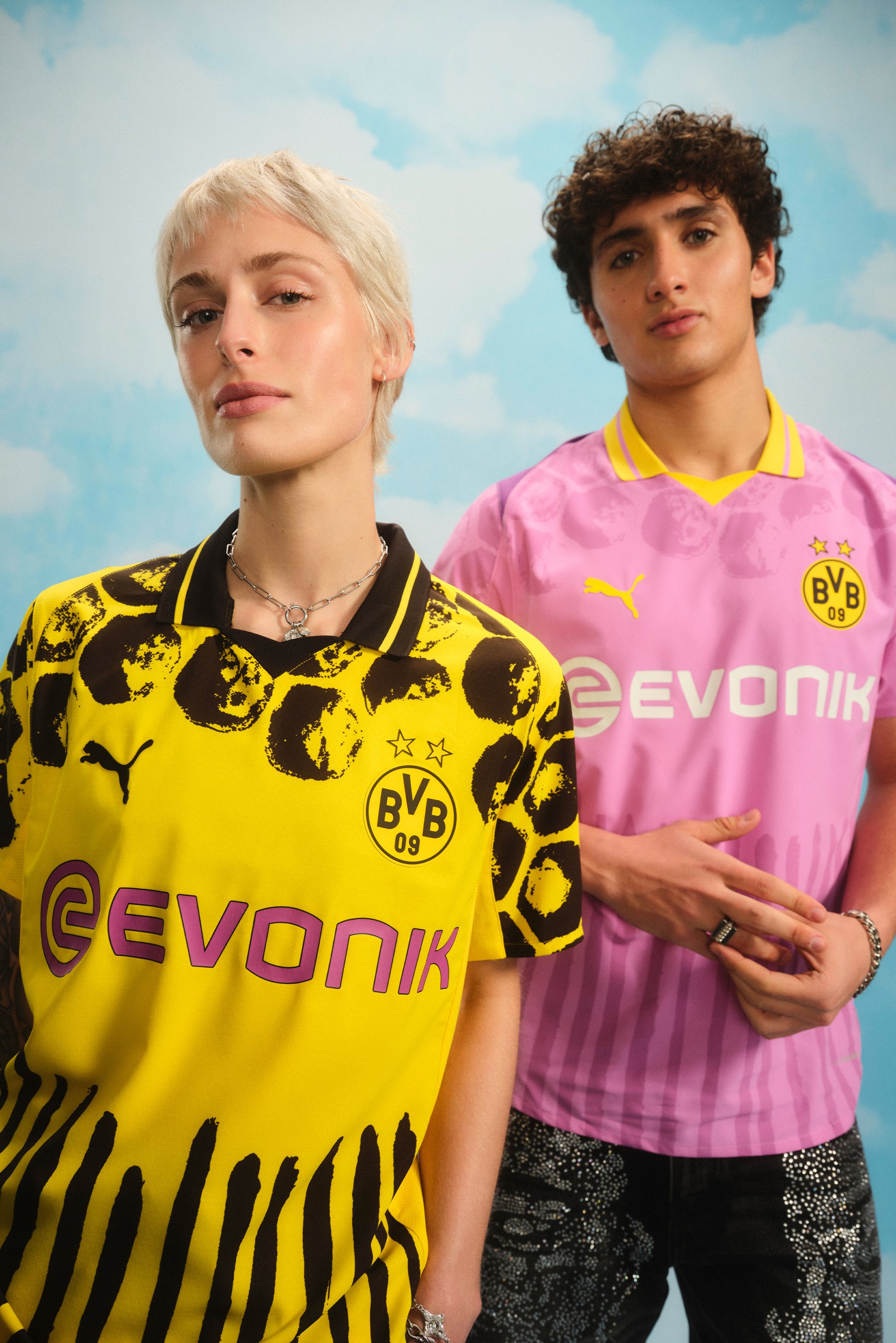 A woman with short blonde hair and a bold yellow patterned shirt stands beside a man in a pink shirt, both posed against a blue sky background.