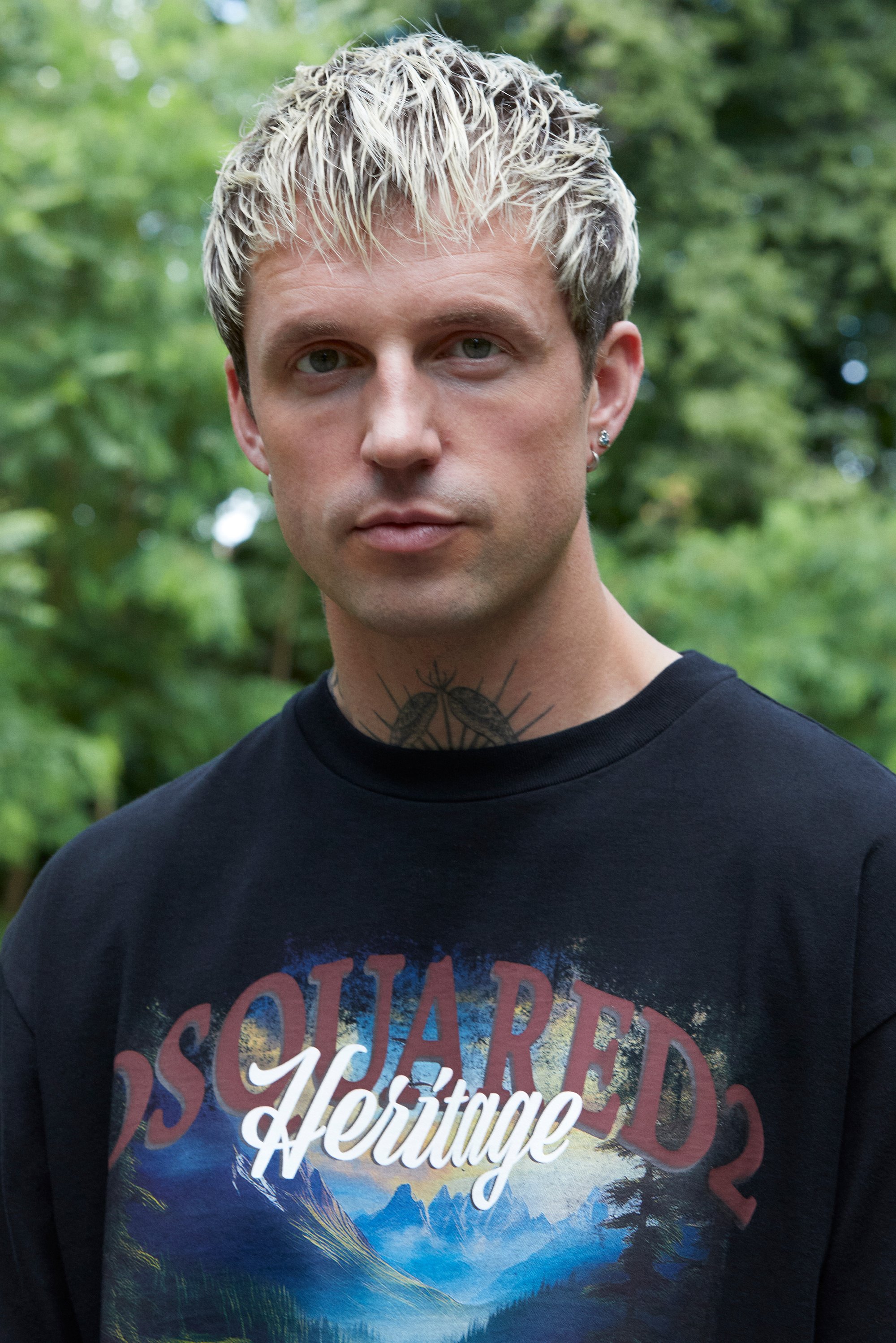 T-shirt nera con grafica "DSQUARED2 Heritage" che presenta un paesaggio montano colorato. Capelli biondi corti e texturizzati con tatuaggi visibili.