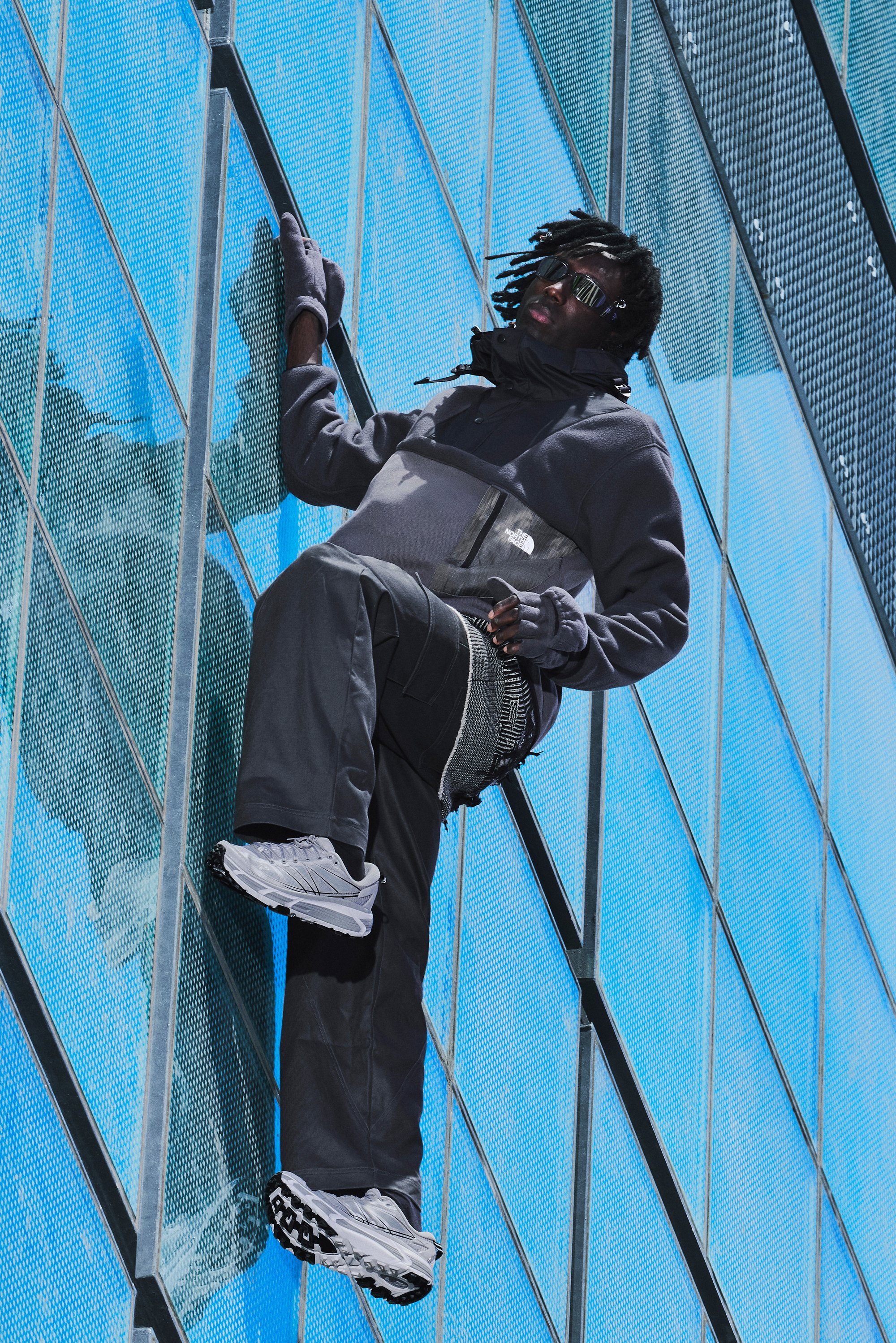 Una persona in abbigliamento sportivo scuro scala un edificio di vetro, mostrando abilità e determinazione contro un luminoso cielo blu.