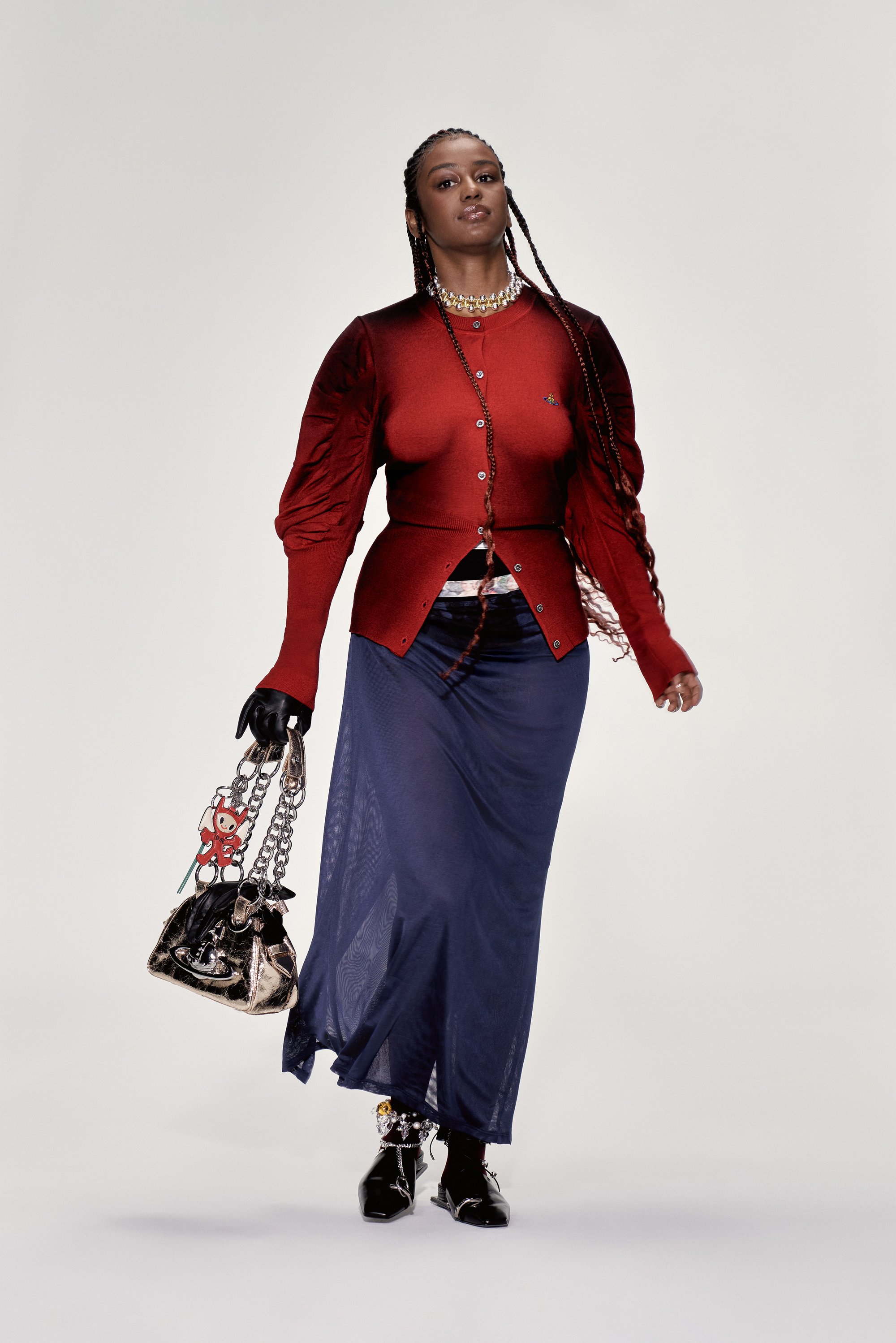Una modelo camina con confianza en una blusa roja y una falda fluida azul marino, llevando un bolso negro adornado con divertidos charms y accesorios.