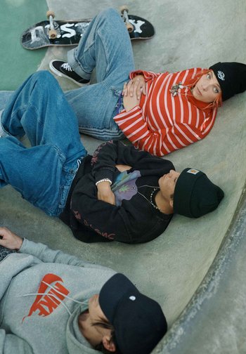 Trois jeunes portant des vêtements décontractés et des chapeaux reposent dans un bol en béton d'un skatepark avec un skateboard à proximité.