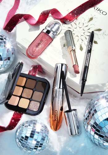 KIKO makeup products on snow: a pink lip gloss, eyeshadow palette with neutral shades, mascara in a silver tube, and various pens. Silver disco balls.