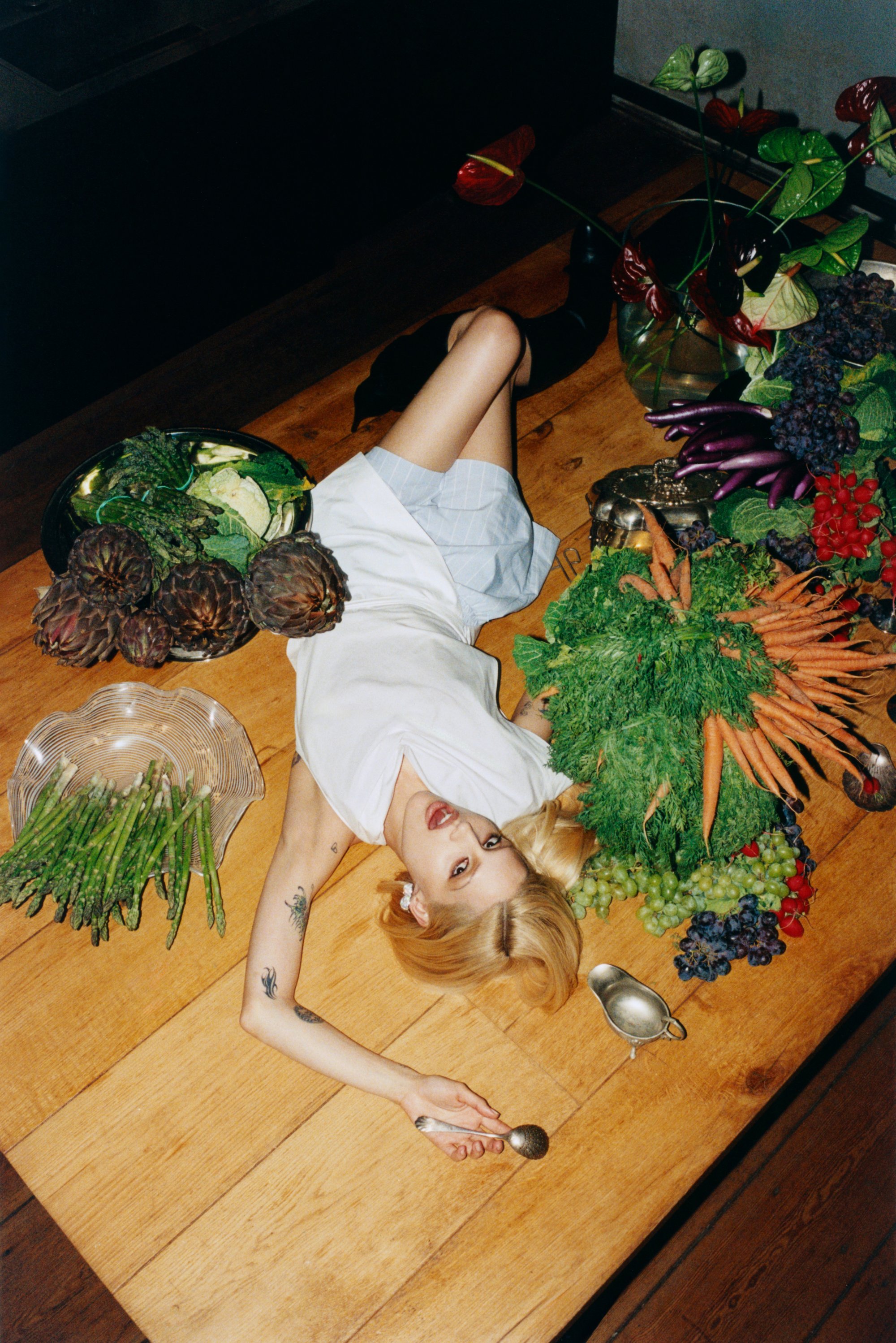 A person lies on a wooden table surrounded by assorted fruits and vegetables, holding a spoon, with a serene expression.