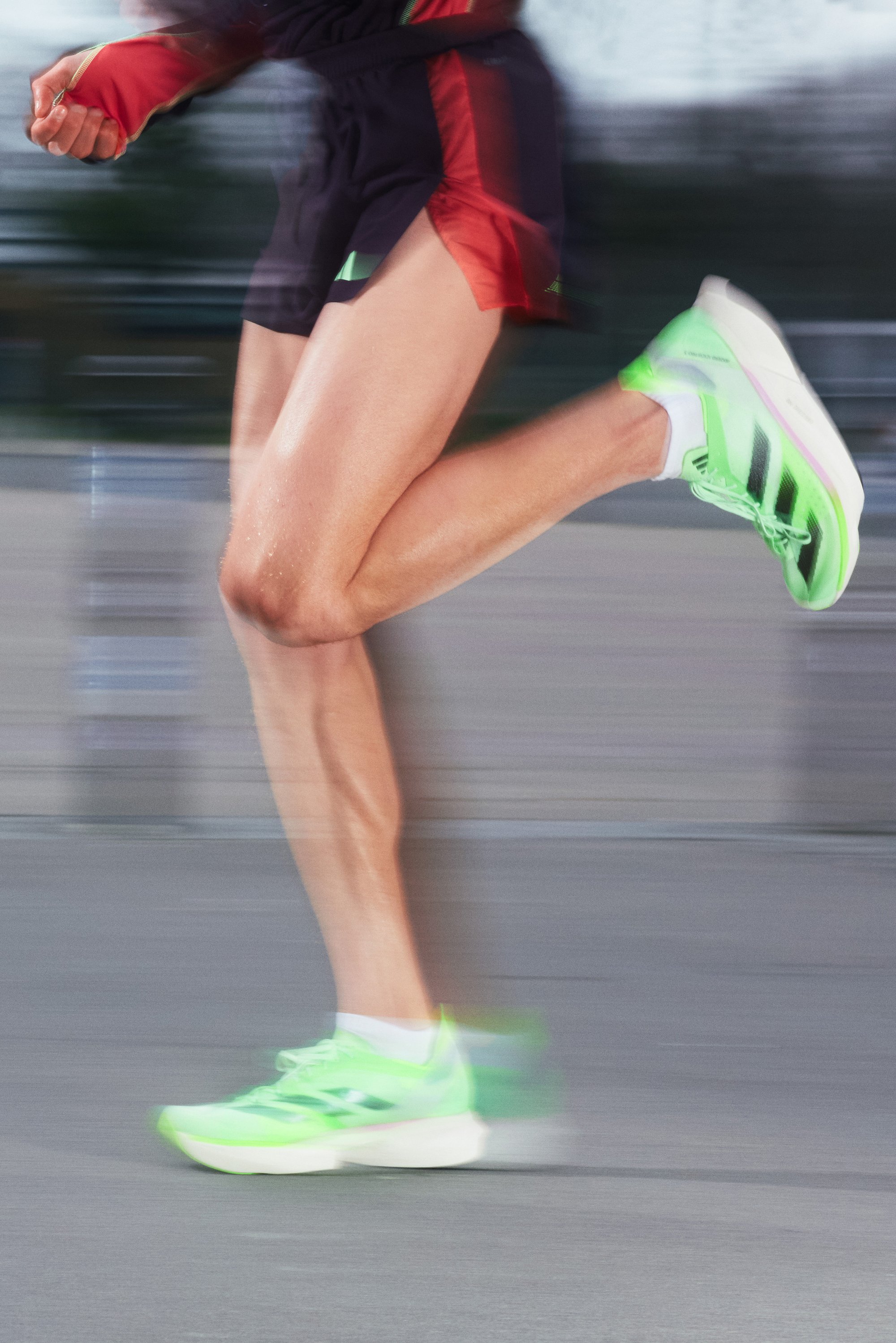 Una pierna en movimiento, vestida con zapatillas de correr de un vibrante verde, parcialmente cubierta por pantalones cortos negros y rojos, que sugieren velocidad y atletismo.