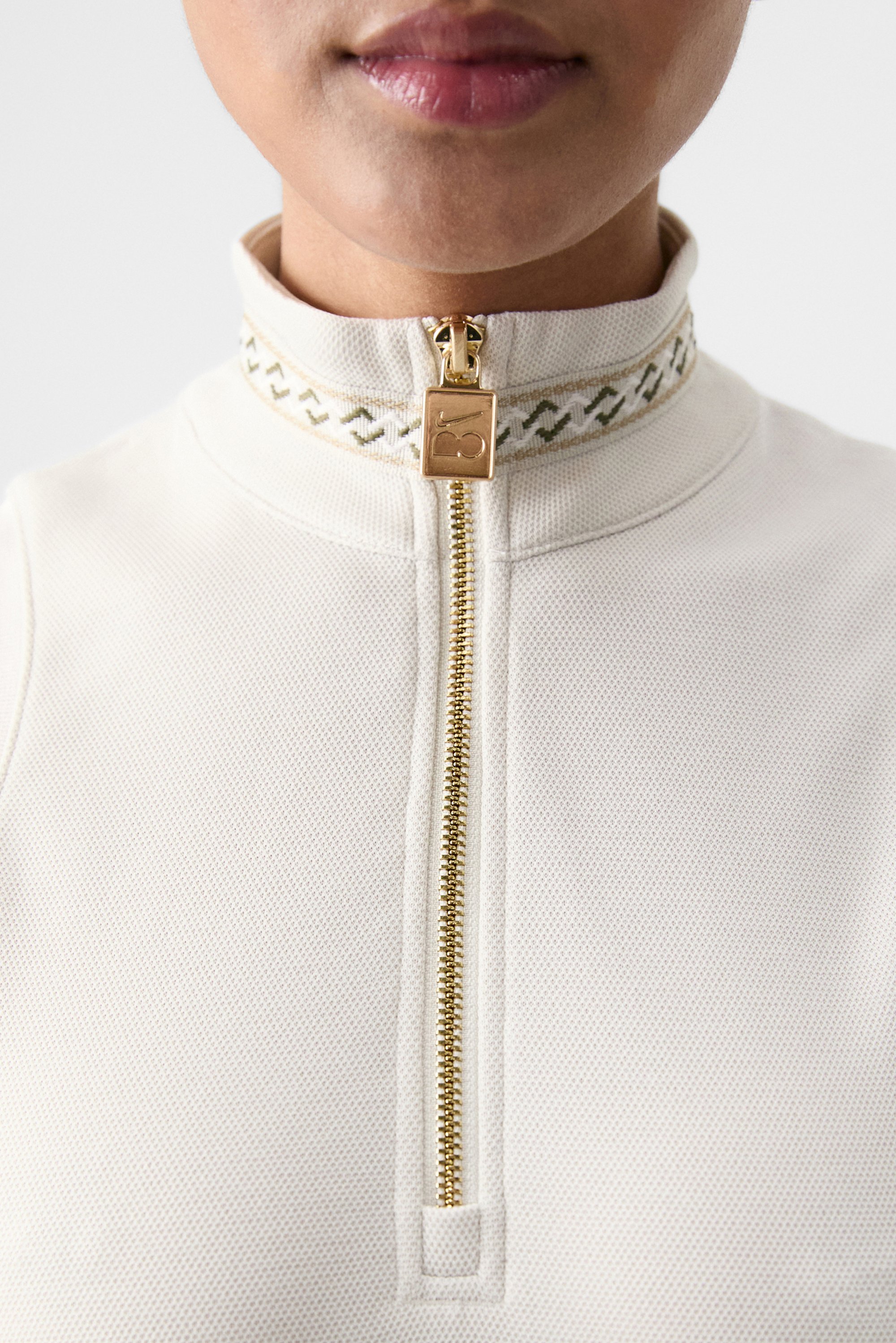 Close-up of a person’s neck featuring a high collar with a gold zipper and decorative pattern, showcasing a stylish, light-colored top.